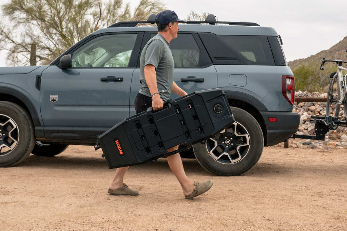 Person carrying a large black box next to a vehicle with bicycles on a bike rack in a desert setting. enlarged image 4
