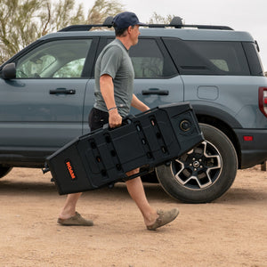 Person carrying a large black box next to a vehicle with bicycles on a bike rack in a desert setting.